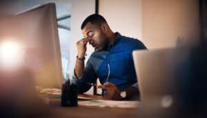 Image of a man sitting at his desk with his hand to his face. He is alone and looks stressed.